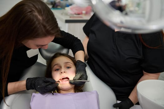 Female dentist doctor in medical protective gloves, holding dental mirror and probe, performing dental treatment and cleaning to a little child girl in dentist chair, curing caries in dentistry clinic