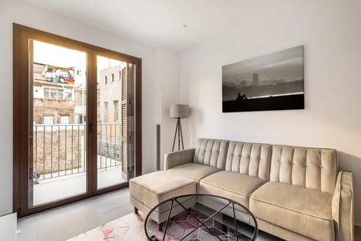 Cozy living room with a corner sofa, a table and a lamp with a picture on the background of a panoramic window with a balcony overlooking the courtyard of the house.