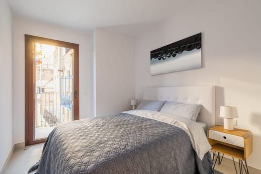 Comfortable double bed in bedroom with a panoramic door-window and a stylish decor in light colors. The concept of a compact cozy apartment for a young family.