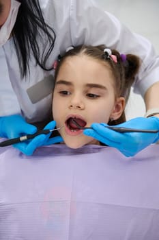 Close-up portrait little child girl in dentist chair, being examined by a doctor dentist. Dental hygienist holding dental tools, performing teeth treatment, curing caries in pediatric dentistry clinic