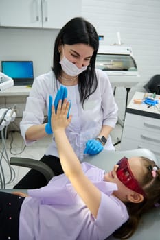Smiling confident female doctor dentist and little patient girl with UV goggles, give high five, sitting in dentist's chair, happy after teeth treatment and regulat dental checkup in dentistry clinic