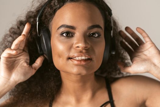 Passion of a music young African-American girl listening her favourite music dancing with hands lifted in headphones wearing black top isolated on cream-grey background, emotionally move, have fun. 