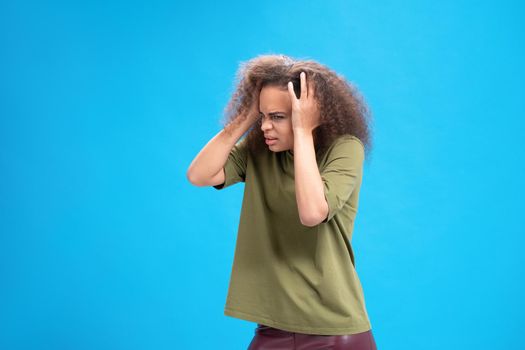 Experiencing migraine or headache African American young woman standing holding head with hands in olive t shirt isolated on blue background. Human emotions concept.