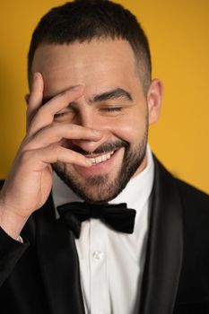 Shy looking down with eye covered with one hand handsome bearded man in tuxedo hide peeps out on closed eyes. Handsome young smiling caucasian man isolated on yellow background. 