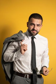 Serious business man dressed in grey suite posing holding his jacket on his shoulder hanging it behind and sunglasses in the other hand looking at camera with a smile isolated on yellow background.