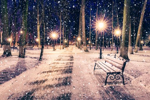 Snowfall in a winter park at night with christmas decorations, lights and  pavement covered with snow. Falling snow.