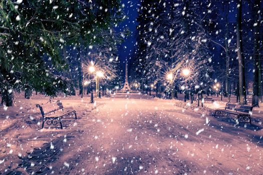 Snowfall in a winter park at night with christmas decorations, lights and  pavement covered with snow. Falling snow.