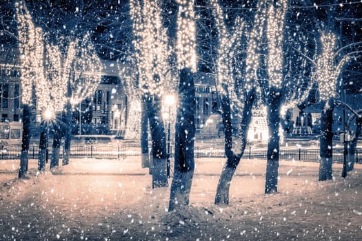 Snowfall in a winter park at night with christmas decorations, lights, pavement covered with snow and trees with garlands.