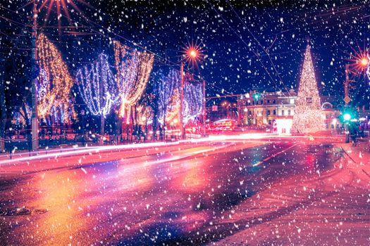 Night city with Christmas decorations, spruce and traces of headlights of moving cars, reflected in the wet asphalt in a snowfall.