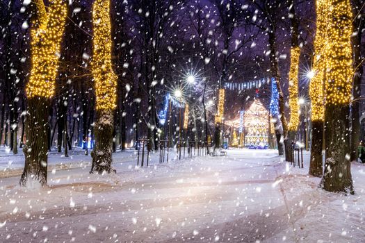 Snowfall in a winter park at night with christmas decorations, lights, pavement covered with snow and trees with garlands.