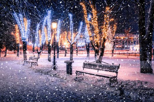 Snowfall in a winter park at night with christmas decorations, lights, pavement covered with snow and trees with garlands.