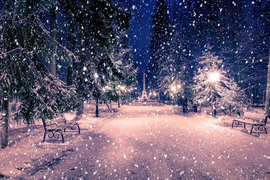 Snowfall in a winter park at night with christmas decorations, lights and  pavement covered with snow. Falling snow.