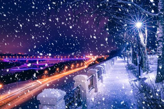 Snowfall in a winter park at night with lanterns, view to road with car motion, pavement and trees covered with snow.