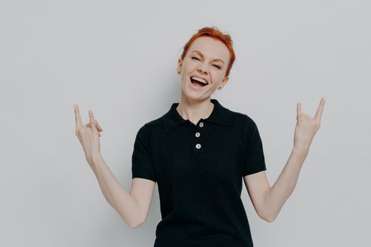 Will rock this party. Energetic crazy young woman with red hair holding hands in rock and roll sign, screaming yelling loudly while standing against grey wall in studio, exclaiming joyfully