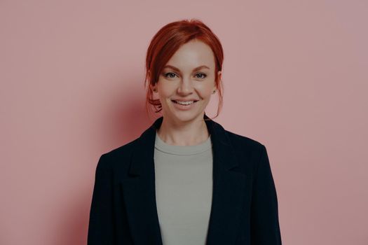 Women beauty concept. Positive young good-looking business woman with red hair looking positively at camera isolated on pink background, expressing happiness and being in good mood