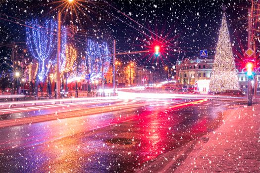 Night city with Christmas decorations, spruce and traces of headlights of moving cars, reflected in the wet asphalt in a snowfall.