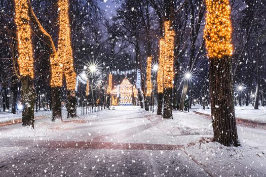 Snowfall in a winter park at night with christmas decorations, lights, pavement covered with snow and trees with garlands.