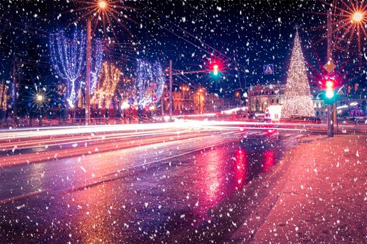 Night city with Christmas decorations, spruce and traces of headlights of moving cars, reflected in the wet asphalt in a snowfall.