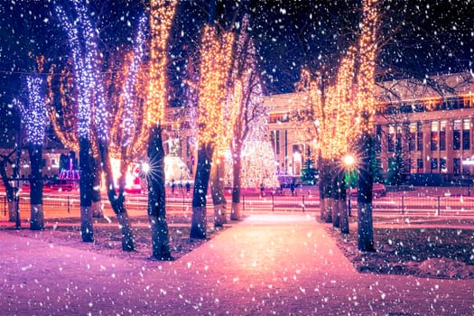 Snowfall in a winter park at night with christmas decorations, lights, pavement covered with snow and trees with garlands.