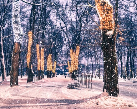 Snowfall in a winter park at night with christmas decorations, lights, pavement covered with snow and trees with garlands.