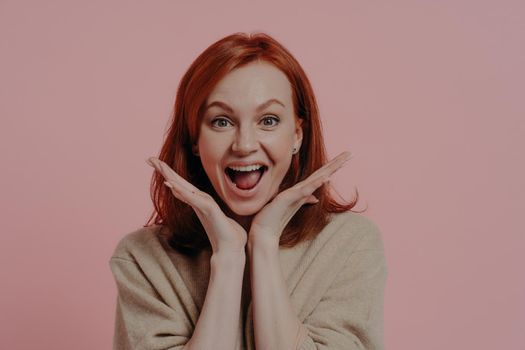 Wow! Portrait of young surprised beautiful ginger woman spreading palms near face and keeping mouth open from excitement, isolated over studio background, dressed in casual clothes. Surprise concept