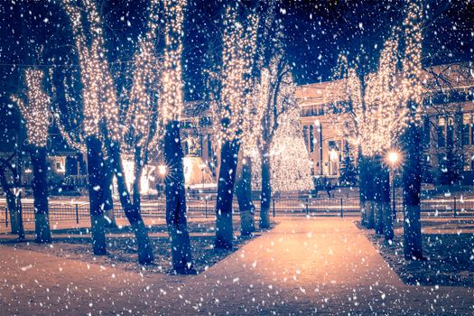 Snowfall in a winter park at night with christmas decorations, lights, pavement covered with snow and trees with garlands.