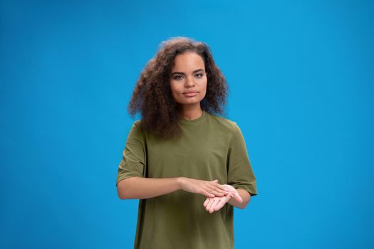 Deaf African American girl shows hand gesture using sign language. Young woman positively looking at camera wearing in an olive t-shirt. Isolated on blue background. Beauty concept. Skin care, hand cream.