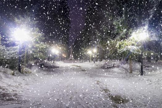 Snowfall in a winter park at night with christmas decorations, lights and  pavement covered with snow. Falling snow.