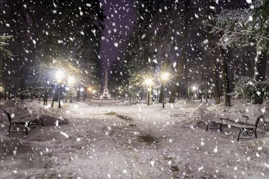 Snowfall in a winter park at night with christmas decorations, lights and  pavement covered with snow. Falling snow.