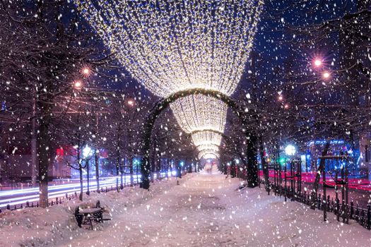 Snowfall in a winter park at night with christmas decorations, lights and  pavement covered with snow. Falling snow.