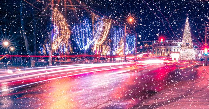 Night city with Christmas decorations, spruce and traces of headlights of moving cars, reflected in the wet asphalt in a snowfall.