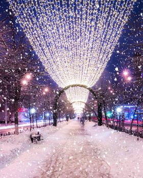 Snowfall in a winter park at night with christmas decorations, lights and  pavement covered with snow. Falling snow.