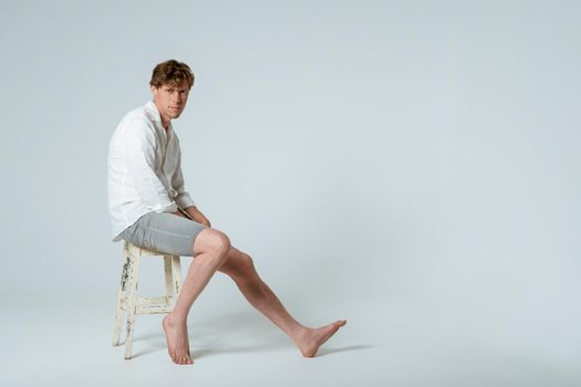 Full length shot of young handsome young man sitting on chair wearing white shirt and grey shorts with one leg out looking at camera isolated on white background. Copy space.