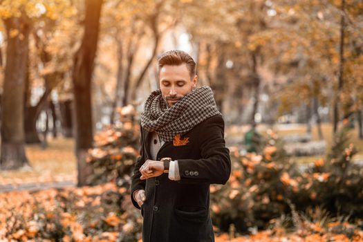 Waiting for his date handsome man looking at his arm watches wearing dark blue coat and scarf. Young freelancer man stands on the street in an autumn coat looking at time on his watches.