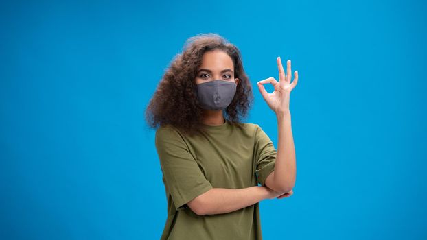 Saying OK young girl with gesture African American woman looking positively at camera wearing olive t-shirt with her arms folded isolated on blue background. Beauty concept.