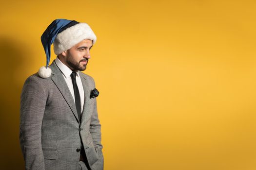 Stylish thoughtful handsome man wearing blue Santa Claus hat and suit, looking at camera isolated on yellow background. Positive emotions, feelings seasonal holidays.