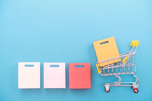 Abstract design element, annual sale, shopping season concept, mini yellow cart with colorful paper bag on pastel blue background, top view, flat lay