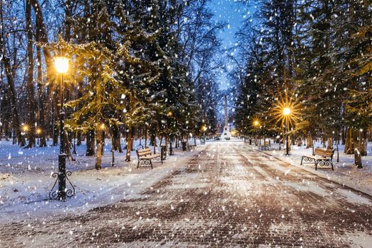 Snowfall in a winter park at night with christmas decorations, lights and  pavement covered with snow. Falling snow.