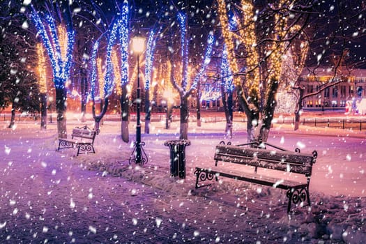 Snowfall in a winter park at night with christmas decorations, lights, pavement covered with snow and trees with garlands.