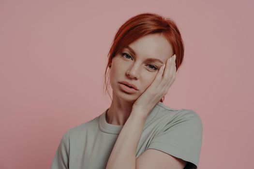 Close up portrait of unhappy tired exhausted ginger woman touching face with hand, suffering from headache or migraine, dressed in casual t-shirt, isolated on pink background
