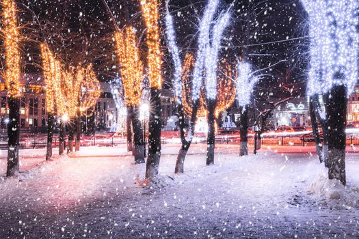 Snowfall in a winter park at night with christmas decorations, lights, pavement covered with snow and trees with garlands.