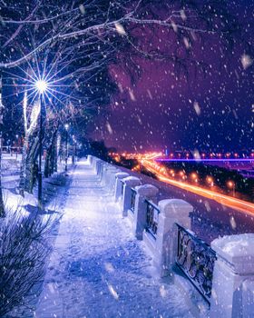 Snowfall in a winter park at night with lanterns, view to road with car motion, pavement and trees covered with snow.