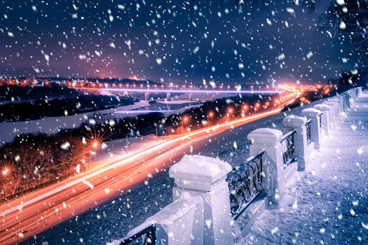 Snowfall in a winter park at night with lanterns, view to road with car motion, pavement and trees covered with snow.