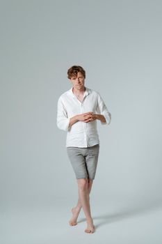 Handsome man in grey jeans shorts and white shirt is standing posing with hands folded and legs crossed in a step and looking at camera. Full length studio shot isolated on white background. 