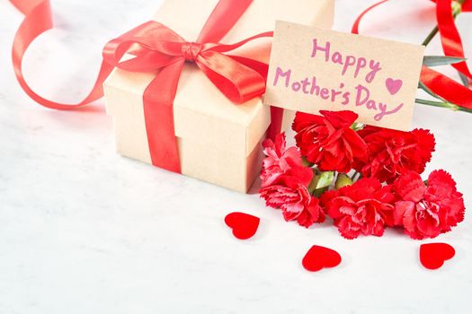 Mother's Day greeting design concept - Beautiful blooming red carnations, gift box with ribbon on white marble table background, close up, copy space.