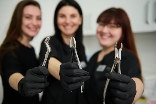 Stainless steel dental pliers, extraction forceps for removing extracting teeth in the hands of blurred smiling female dentists doctors, maxillofacial surgeons, orthodontists in black medical uniform