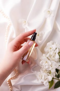 A woman's hand holds a bottle of perfume over a background with flowers on a white satin fabric.