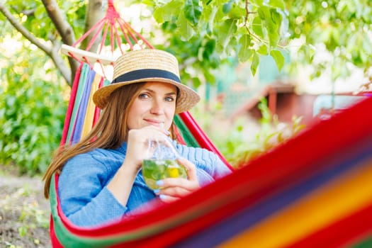 Young woman drinking a cocktail while lying in comfortable hammock at green garden.