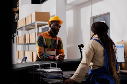 Warehouse workers standing at counter desk discussing shipment management. Two african american post office employees talking about package delivery at checkout in storage room