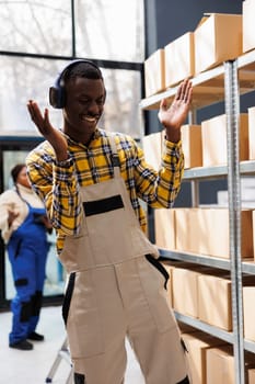 Happy african american man enjoying music in warehouse at work, dancing and clapping hands. Optimistic young storehouse worker wearing headphones moving in parcels storage room aisle